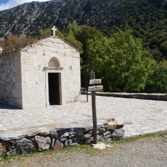 Small stone chapel with a cross on the roof, set beside a mountain slope and trees in South Crete