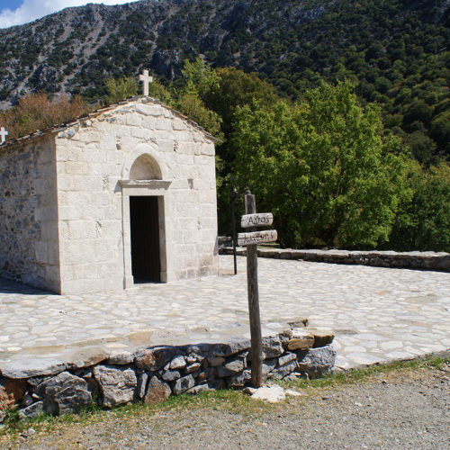 Small stone chapel with a cross on the roof, set beside a mountain slope and trees in South Crete
