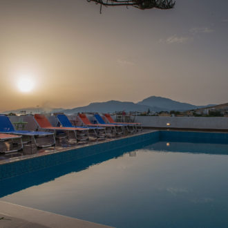 Poolside loungers beside a calm swimming pool at sunset, with mountains and village rooftops in the distance