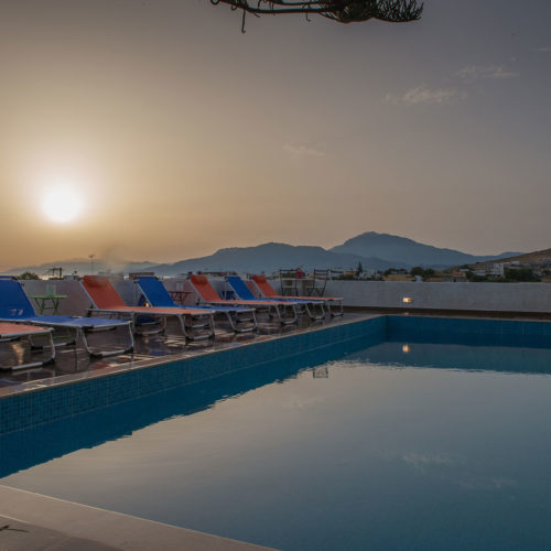Poolside loungers beside a calm swimming pool at sunset, with mountains and village rooftops in the distance