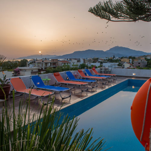 Pool with orange and blue loungers at sunset, overlooking Kalamaki, Crete and distant hills