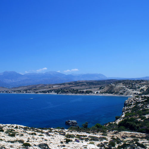 Wide coastal bay with deep blue water, rocky cliffs, and distant mountains under a clear sky