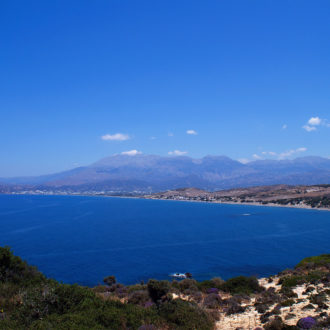 Wide coastal view of a blue bay, sandy shoreline, and distant mountains under a clear sky