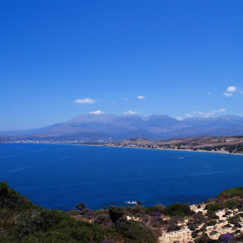 Wide coastal view of a blue bay, sandy shoreline, and distant mountains under a clear sky
