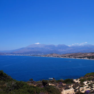 Wide coastal view of a blue bay with mountains and rocky shoreline under a clear sky