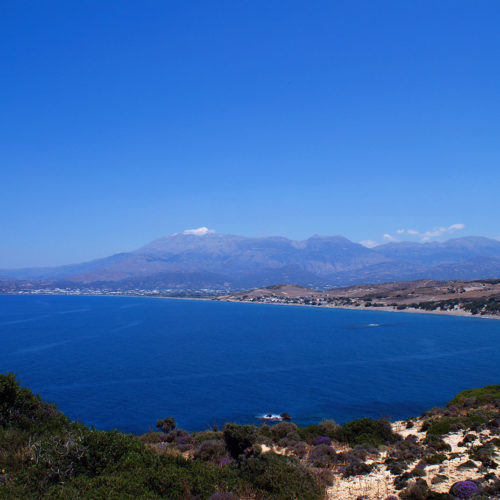 Wide coastal view of a blue bay with mountains and rocky shoreline under a clear sky