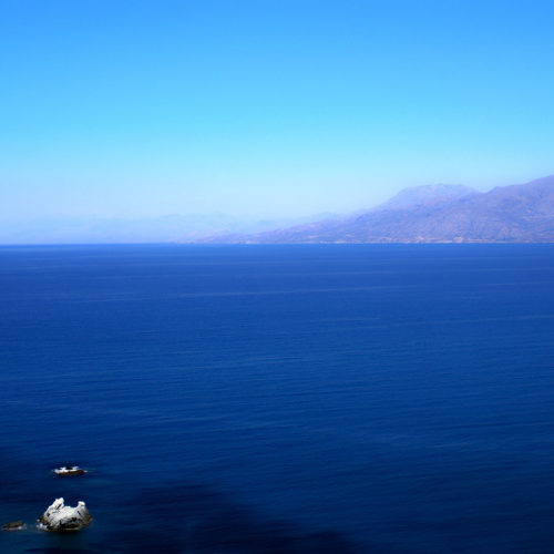 Wide view of calm blue sea with distant mountains and a small rocky outcrop